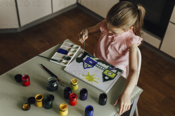 A child, dressed in a pink top, is intently painting a vibrant scene of houses and a sun using