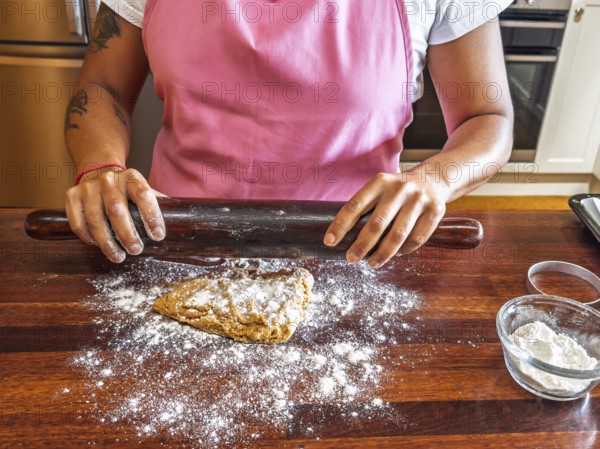 A woman in a pink apron uses a dark rolling pin to flatten dough on a floured wooden counter. A