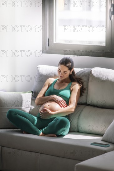 A pregnant woman peacefully sitting on a sofa in a sunlit room, dressed in a comfortable green yoga