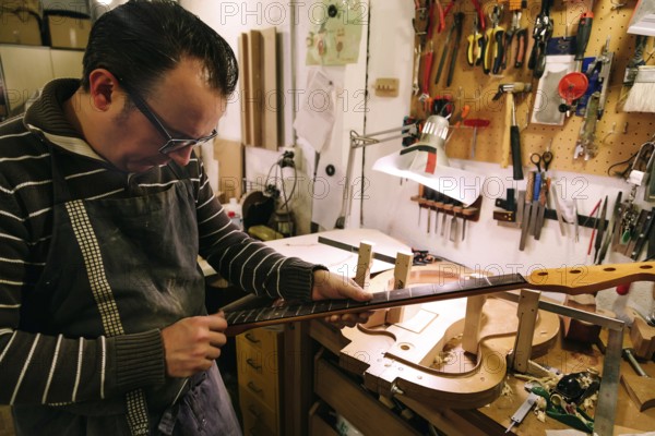A luthier meticulously crafts a bass guitar neck in a workshop filled with tools. The focus is on