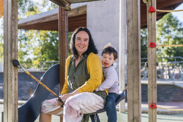 A joyful mother and child with Down syndrome are sitting together on a playground swing on a sunny