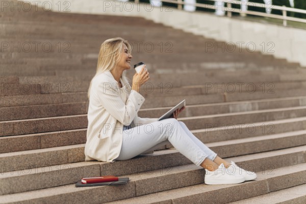 A businesswoman sits on outdoor steps with a tablet, enjoying a coffee. Dressed casually in white