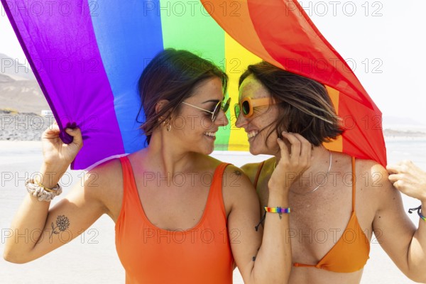 A joyful lesbian couple shares a moment under a colorful rainbow flag on a sunny beach. Both are