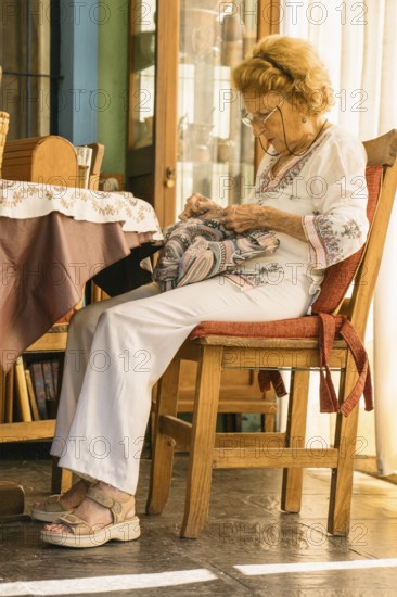 Elderly woman wearing glasses and white clothes is focused on sewing a colorful fabric while