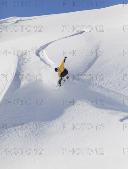 A snowboarder in a yellow jacket executes a jump on a snowy mountain slope. The clear blue sky and