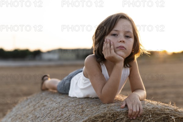 Child in casual attire lies on a hay bale during sunset, reflecting a calm and serene mood. The