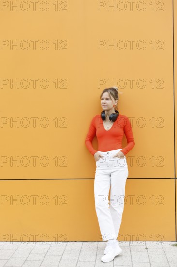 A fashionable woman leans against a striking orange wall in Warsaw, wearing bright attire with