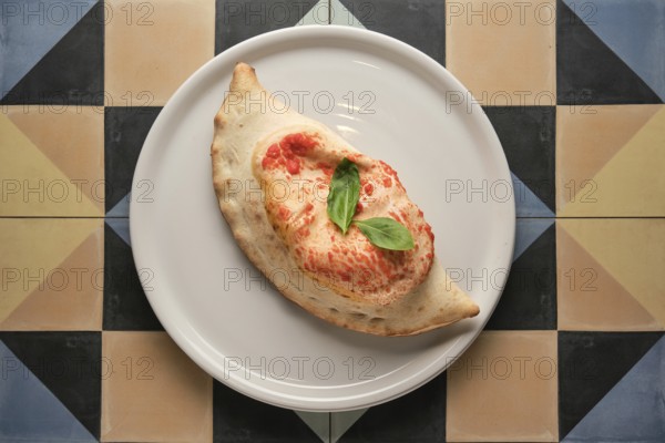 Freshly baked calzone garnished with basil leaves, served on a white plate over a vibrant geometric
