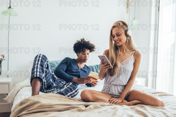 A couple enjoys a relaxed morning on a cozy bed, one listens to music on headphones, the other