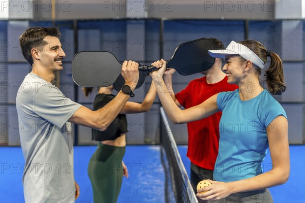 Four friends on a blue pickleball court paddle to paddle over the net, smiling and high fiving