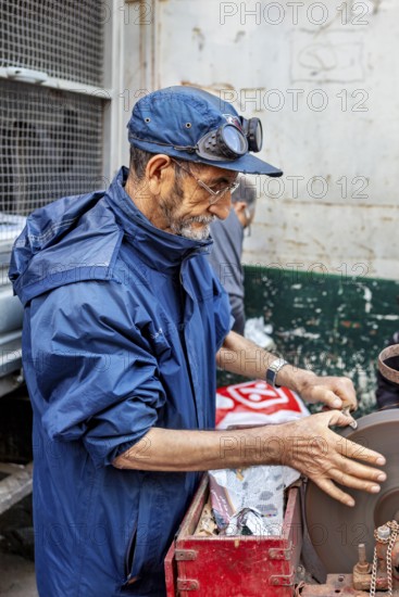 A man working on a machine in a workshop, wearing a blue jacket and a cap, A man sharpening knives