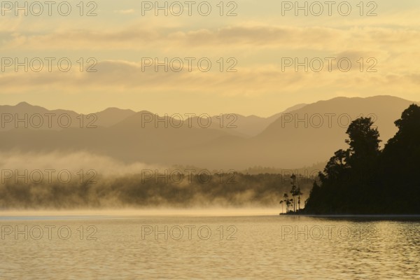 Mist drifts over a calm lake with mountains in the background at sunrise, Lake Mahinapua, Ruatapu,