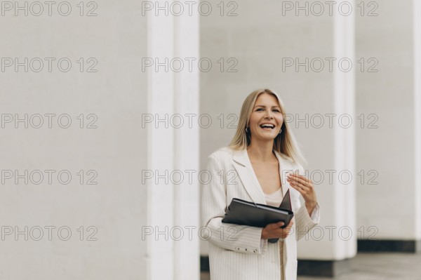 A businesswoman in a white suit smiles joyfully while holding a folder. She stands in front of
