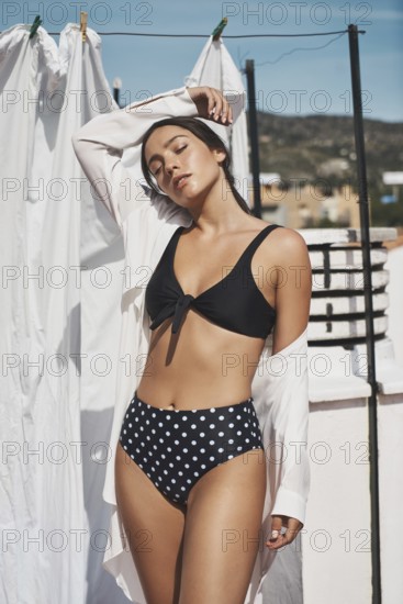 A young woman in a black polka-dot bikini and white shirt enjoys the summer sun on a rooftop The
