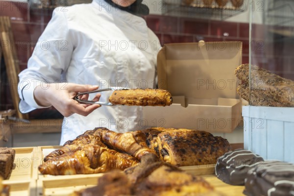 Bakery worker in uniform uses tongs to place a warm pain au chocolat into a paper takeaway box amid
