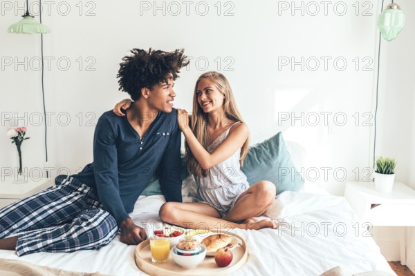 A young couple sits cheerfully on a bed, sharing a breakfast tray filled with fruit, juice, and