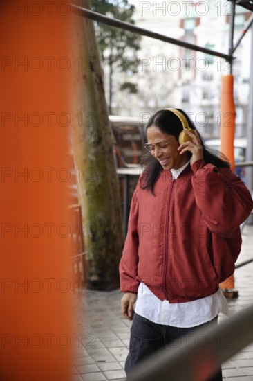 A man with long hair and glasses enjoys music through yellow headphones while walking outdoors in a