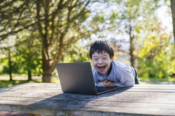 A cheerful child with Down syndrome is smiling joyfully at a laptop while sitting at a wooden table