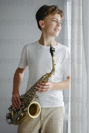 A young saxophonist in a white shirt looks out the window, standing in soft sunlight. The relaxed