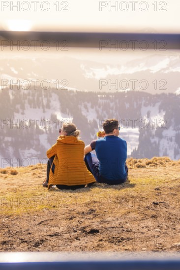 Couple sitting on a mountain peak with a view of the sunny winter valley, Hochgrat Mountains,