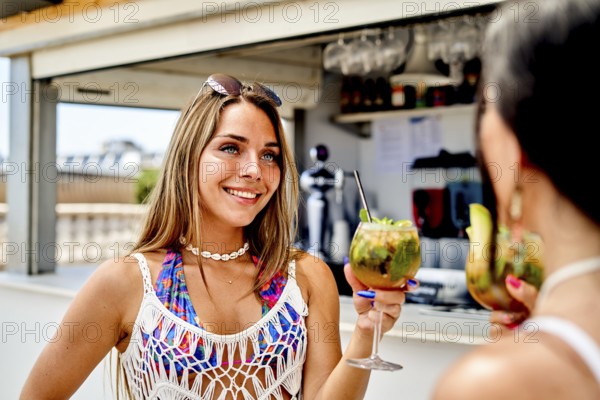 Two friends are at a vibrant beach bar during a sunny summer day, holding colorful cocktails and