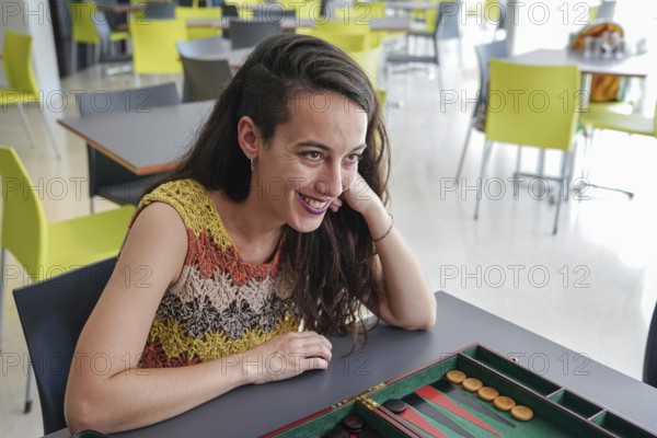A woman sits at a table in a modern cafe, smiling and engaged, while playing a game of backgammon
