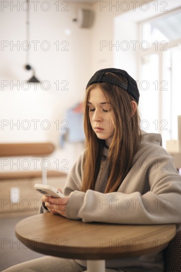 A teenage girl wearing a hoodie and cap sits at a small table, engrossed in her smartphone. The