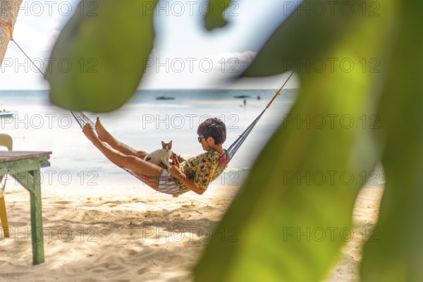A woman and a cat enjoy a serene moment in a hammock on a sandy beach, surrounded by lush green