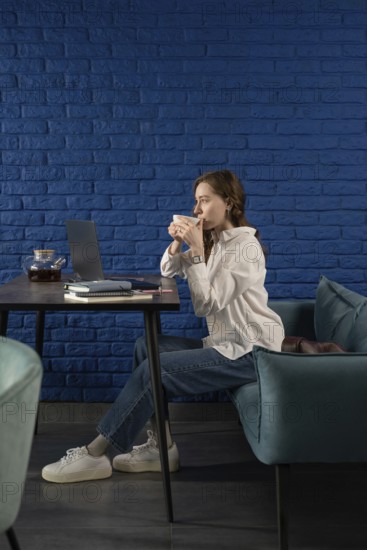 A woman in casual attire sips tea, seated at a table with a laptop, against a vibrant blue brick