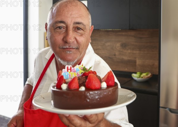 A cheerful senior man in a red apron holds a chocolate cake adorned with strawberries and a Happy