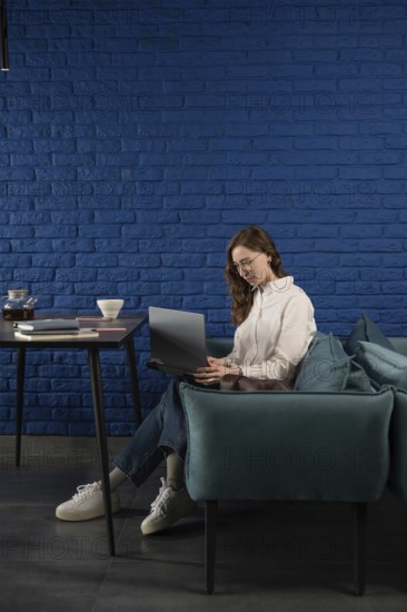 A woman in casual attire works on a laptop, seated on a teal sofa against a blue brick wall. A