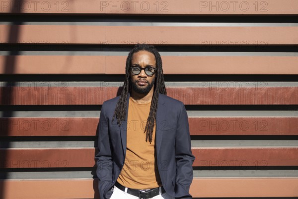 Stylish cuban business man with dreadlocks in sunglasses and casual blazer stands against modern