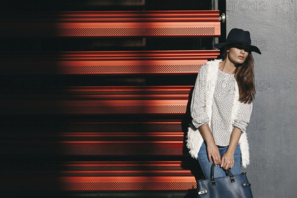 A fashionable woman in a wide-brimmed hat and stylish clothing poses with a handbag against a red