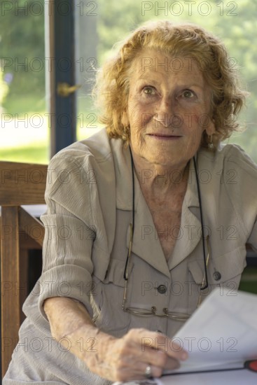 Portrait of a senior woman sitting at a table on a sunny veranda, holding and reading a book,