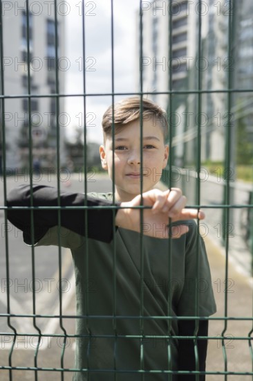 A young boy in a dark shirt stands pensively behind a green wire fence, with urban high-rise