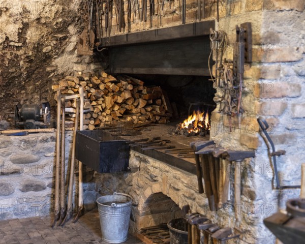 A rustic blacksmith workshop featuring a burning forge, neatly stacked firewood, and an array of