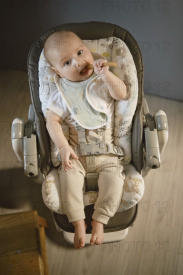 A baby is sitting in a high chair, holding a spoon and looking at something. The room has soft