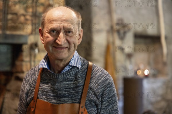 A skilled blacksmith wearing a leather apron smiles warmly in his rustic workshop. The background