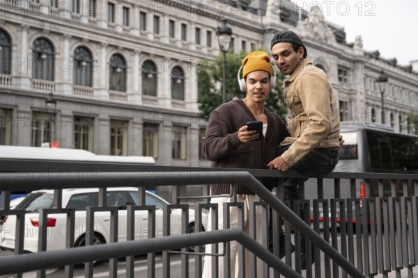 A gay couple in casual attire shares a tender moment on a Madrid street, leaning on a railing and