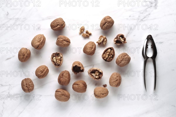 Whole and cracked walnuts scattered on a marble surface, next to a shiny metal nutcracker. The