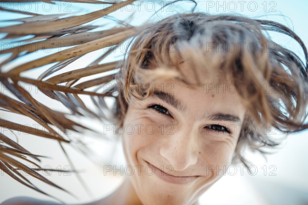 A joyful teenage boy smiles under the dappled shade of a palm tree, his hair tousled by a gentle