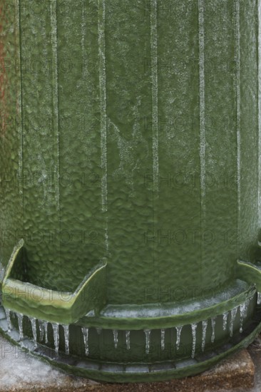 Upside down green PVC and rubber thrash can covered with thick sheet of ice and hanging icicles