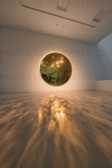 A round mirror on the wall casts warm light reflections onto the floor in an art space, Schauwerk