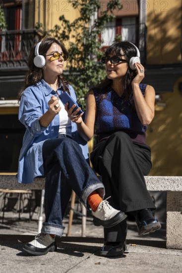 A multiethnic lesbian couple sits on a bench outdoors, wearing headphones and sharing a joyful