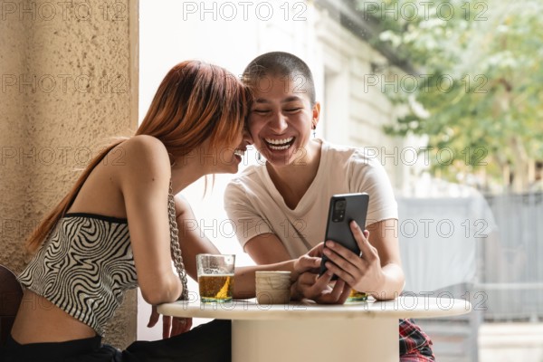 Two lesbian women share a joyful moment at a cafe, laughing and looking at a smartphone. They sit