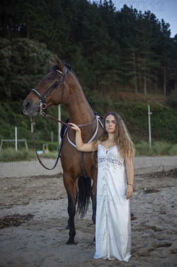 A young woman in a flowing white dress stands beside her horse on a tranquil beach. The scene