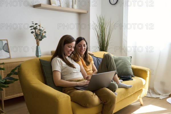 A lesbian couple sits comfortably on a yellow couch, smiling and using a laptop together in a cozy