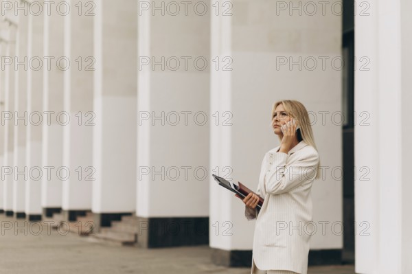 A businesswoman in a beige suit talks on her phone while standing outside an office building with
