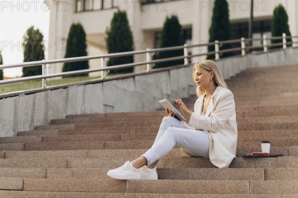 A businesswoman in casual attire sits on outdoor steps with a digital tablet, concentrating on her