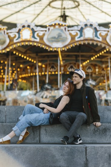 A couple sits on stone steps, the woman leaning on the man, in front of a brightly lit carousel in